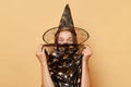 Surprised young woman wearing witch costume and carnival cone hat celebrating halloween isolated over beige background covering Royalty Free Stock Photo