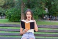 Surprised young woman with widely open yeas and mouth and a hand on cheek is reading a book outdoors