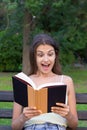 Surprised young woman with widely open yeas and mouth is reading a book outdoors