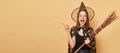 Surprised young woman wizard wearing witch costume holding in hand broom  over beige background pointing away at Royalty Free Stock Photo