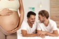 Surrogacy concept. Young pregnant woman and blurred view of happy gay couple indoors
