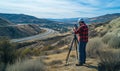 Surveyor using equipment in mountainous landscape, capturing data for project, highway visible in background, clear blue Royalty Free Stock Photo