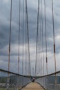 suspended pedestrian bridge with metal vertical ropes, cables and wooden path against the backdrop of cloudy sky, geometric
