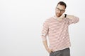 Suspicious serious handsome male model in glasses, holding hand near ear and listening to watch, standing over gray wall Royalty Free Stock Photo