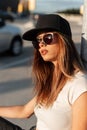 Swag modern young woman hipster in fashion casual t-shirt in trendy baseball black cap in stylish sunglasses sit on asphalt at Royalty Free Stock Photo