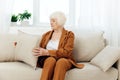 a sweet, amiable elderly woman in a brown suit is sitting on a beige wide sofa, adjusting a decorative pillow with her