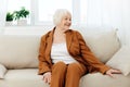 a sweet, amiable elderly woman in a brown suit is sitting on a beige wide sofa, smiling pleasantly while in the