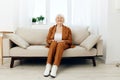 a sweet, amiable elderly woman in a brown suit is sitting on a beige wide sofa, smiling pleasantly while in the