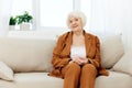 a sweet, amiable elderly woman in a brown suit is sitting on a beige wide sofa, smiling pleasantly while in the