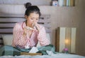 Sweet Asian Korean girl in pajamas covered with blanket sick suffering cold and flu taking temperature with thermometer in bed