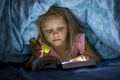 Sweet beautiful and pretty little blond girl 6 to 8 years old under bed covers reading book in the dark at night with torch light Royalty Free Stock Photo
