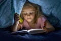 Sweet beautiful and pretty little blond girl 6 to 8 years old under bed covers reading book in the dark at night with torch light Royalty Free Stock Photo