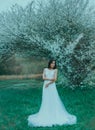 Sweet charming young woman with long black hair and emerald eyes in front of blooming white magnolias stands in amazing