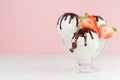 Sweet creamy ice cream in elegant glass bowl with ripe strawberry slices, chocolate sauce on white wood table and pastel pink wall