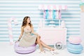 Sweet lifestyle, cheerful mood of joyful pretty young woman in dress sitting on big macaron among sweets on striped