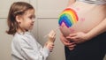 Sweet little daugter painting pregnant belly her mother. Pregnant mom and her child having fun together at home. Family, healthy