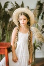A sweet little girl with long blond hair in a white sarafan and a black hat in a rattan chair