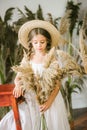 A sweet little girl with long blond hair in a white sarafan and a straw hat in a rattan chair