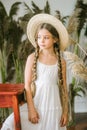 A sweet little girl with long blond hair in a white sarafan and a straw hat in a rattan chair