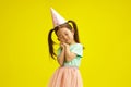 Sweet little girl in party hat dreamly closed eyes, cute placed palms of hands in front of herself face, looks like as Royalty Free Stock Photo