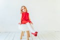 Sweet little girl in red dress sitting on chair against white wall at home, relaxing in white bright living room indoors. Royalty Free Stock Photo