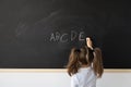 Sweet little girl at school in a lesson. A child stands in front of a black board. She writes in English the letters of