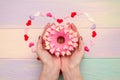 Sweet pink donut with icing and decoration with hearts on rainbow wooden background in female hands