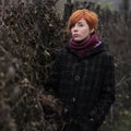 Sweet red-haired girl in a black coat and purple knitted scarf is standing by the fence overgrown with grapevine or ivy, girl or w Royalty Free Stock Photo