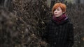 Sweet red-haired girl in a black coat and purple knitted scarf is standing by the fence overgrown with grapevine or ivy, girl or w Royalty Free Stock Photo