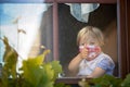 Sweet toddler boy, wearing medical mask, hugging teddy bear, also with mask, looking sadly out of the window, during coronavirus Royalty Free Stock Photo