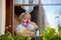 Sweet toddler boy, wearing medical mask, hugging teddy bear, also with mask, looking sadly out of the window, during coronavirus Royalty Free Stock Photo