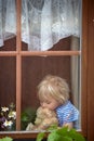 Sweet toddler boy, wearing medical mask, hugging teddy bear, also with mask, looking sadly out of the window, during coronavirus Royalty Free Stock Photo