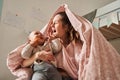 Adorable happy young mother sitting under the blanket at the bedroom with her newborn son