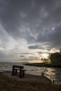 A swooden sitting bench on a lake shore at sunset, beneath a moody, cloudy sky