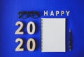 Symbol Happy new year 2020 year, with plywood number 2020, notebook, black vintage glasses and a pencil on blue paper background Royalty Free Stock Photo