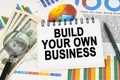 On the table are business charts, dollars and a notepad with the inscription - Build Your Own Business