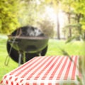 Table with checkered tabletop and blurred grill with smoke on grass and garden background. Royalty Free Stock Photo