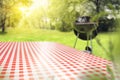 Table with checkered tabletop and blurred grill with smoke on grass and garden background.