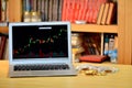 On the table golden bitcoins and laptop with stock exchange graph on screen, blurred shelf with books in the background