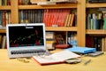 On the table golden bitcoins, notebook, eyeglases and laptop with stock exchange graph on screen, blurred shelf with books in the