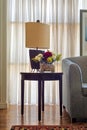 Table lamp and colorful flowers planter on small dark brown wooden table on background of big window with white sheer curtains