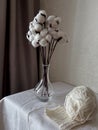 on the table there is a vase with cotton flowers and there is a skein of white wool with a knitted square started