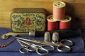 OLD PIN BOX WITH REELS OF THREAD, THIMBLES, SCISSORS, GLASSHEAD PINS AND TRACING WHEEL ON A PIECE OF DENIM MATERIAL