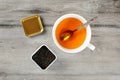 Table top view on cup of hot amber tea with spoon, and caddy full of black dried loose leaves, placed on gray wood desk.