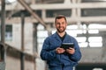 With tablet in hands. Factory worker in blue uniform is indoors