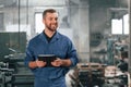 With tablet in hands. Factory worker in blue uniform is indoors