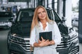 With tablet in hands. Woman in formal clothes is in the car dealership