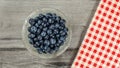 Tabletop view, small glass bowl of blueberries, red checkered gingham tablecloth next to it on gray wood desk