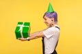 Take birthday gift! Side view of positive cute hipster girl wearing funny party cone, giving present box. studio shot