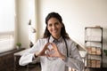 Smiling latin female cardiologist look at camera show finger heart